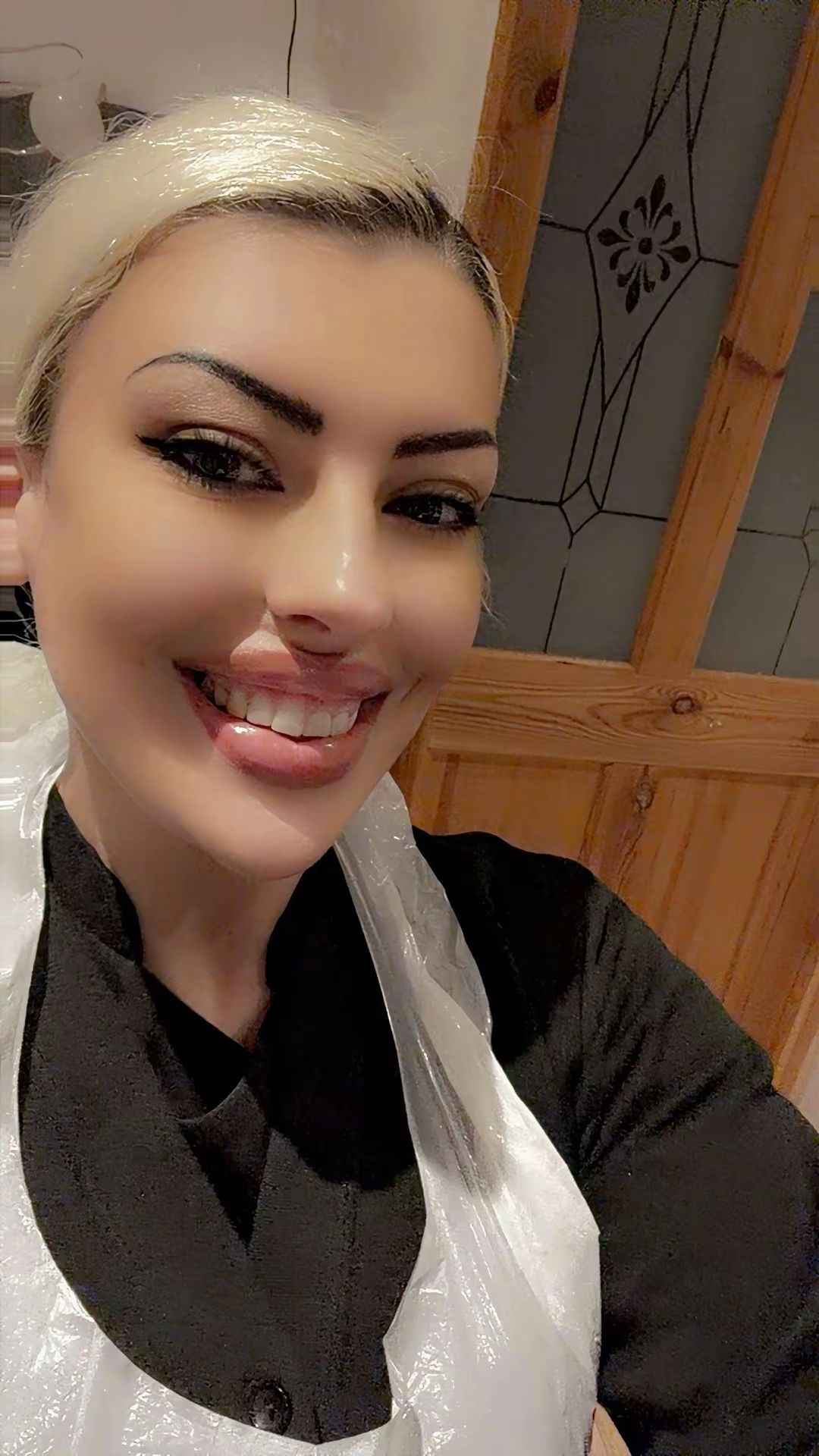 Ffion smiling warmly at the camera in a selfie. She has platinum blonde hair pulled back, bold dark eye makeup with cat-eye liner, and glossy lips. She is wearing a black tunic with a clear plastic apron over the top, standing in what looks like her treatment room with a wooden-framed stained glass door panel visible behind her.